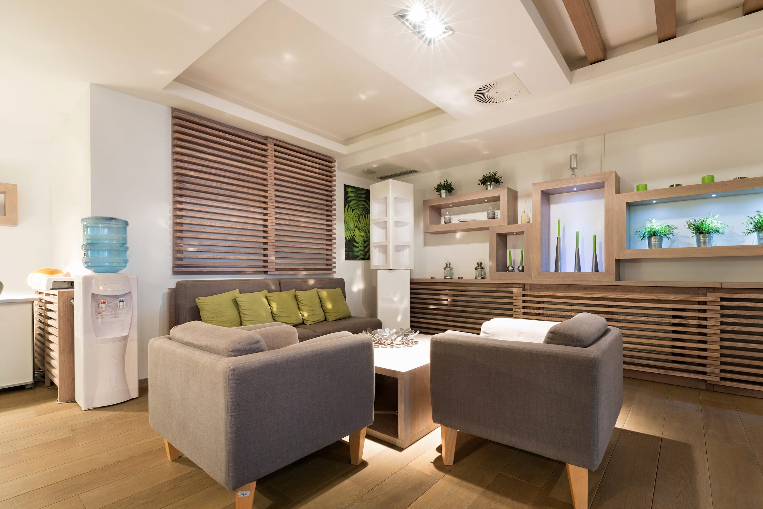 A modern lounge area featuring two gray armchairs with green cushions, a small coffee table, and wooden flooring. A water cooler is on the left, and a wall shelf with plants is seen in the background, along with ambient lighting.