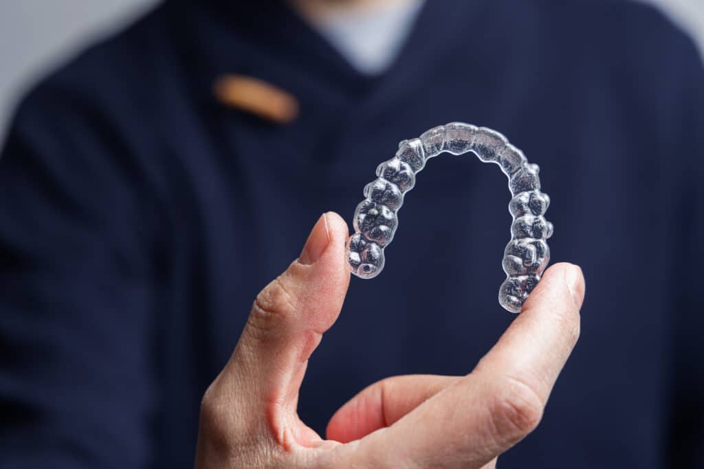 A person holding an invisible aligner, a clear orthodontic device, between their fingers. The aligner is in the shape of an arch and has small buttons. The background is slightly blurred, focusing on the aligner.