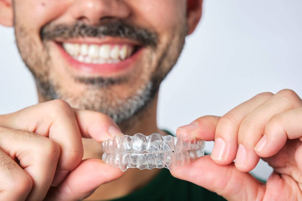 A man smiling broadly while holding a clear dental aligner in both hands. He has a short beard and is wearing a green shirt. The background is a light gray, emphasizing his expression and the aligner.