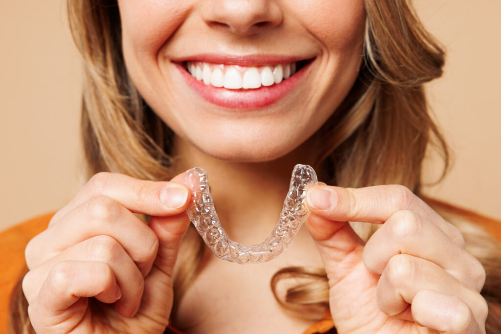 A woman smiling while holding a clear dental aligner tray in front of her. She has long brown hair and is wearing a mustard-colored shirt. The background is a soft beige color, emphasizing her smile and the aligner.