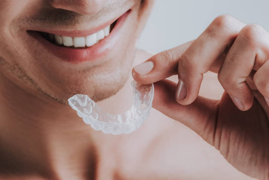 A close-up image of a man smiling while holding a clear orthodontic aligner near his mouth. He shows straight, white teeth and has stubble on his chin. The background is neutral and softly blurred, emphasizing the aligner.