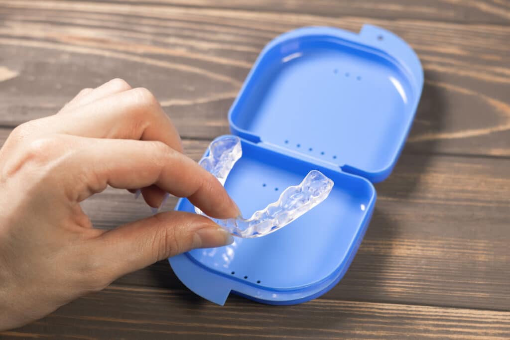 A person's hand holds a clear dental retainer above a blue storage case that is open. The retainer is visible, with its curved shape, resting near the bottom of the case. The background is a wooden surface.