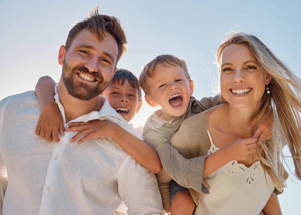A smiling family of four posing outdoors. A man wears a white shirt, carrying a young boy on his back. Another boy is in the middle, smiling widely. A woman beside them, dressed in a beige top, is gently holding a boy's shoulders, all against a bright sky.