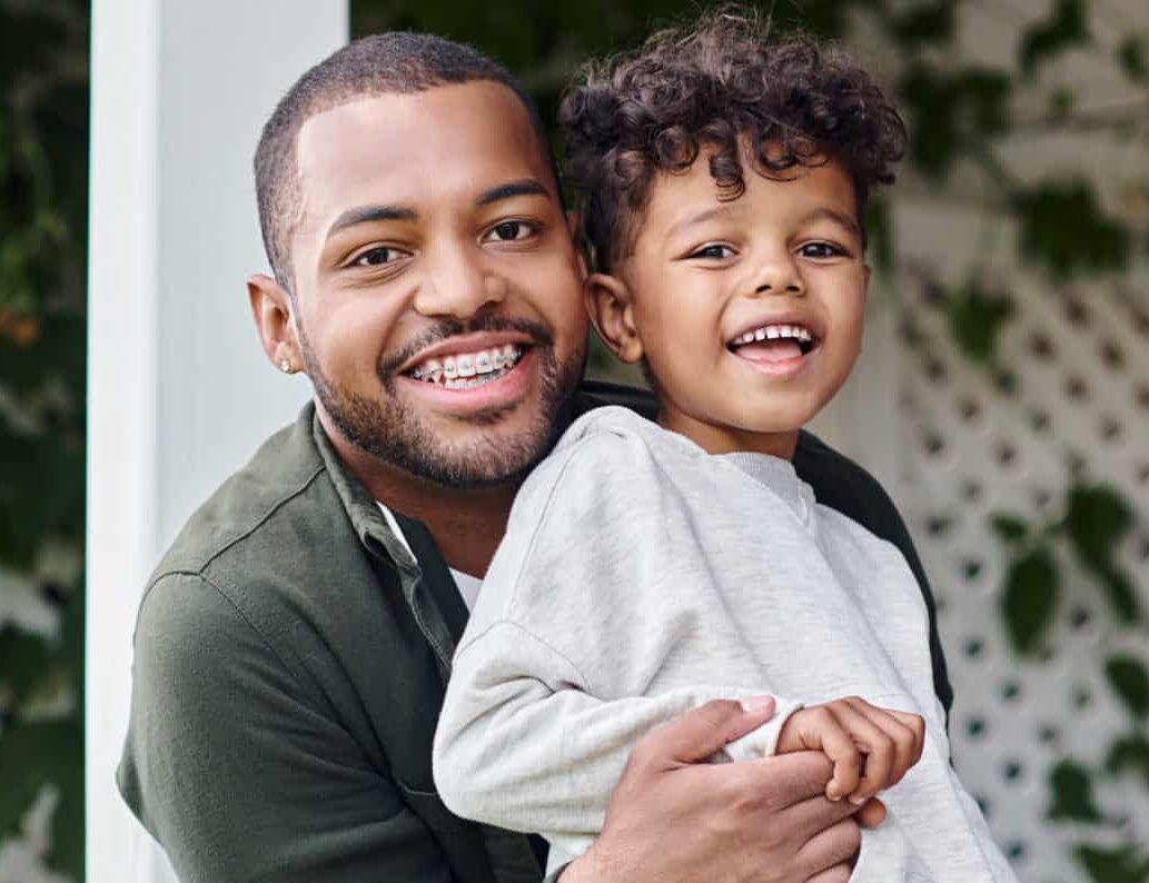 A smiling man holding a child in his arms outside, with green plants and a white lattice background. The child, wearing a light gray outfit, looks happy as they both face the camera. The setting suggests a warm, outdoor environment.
