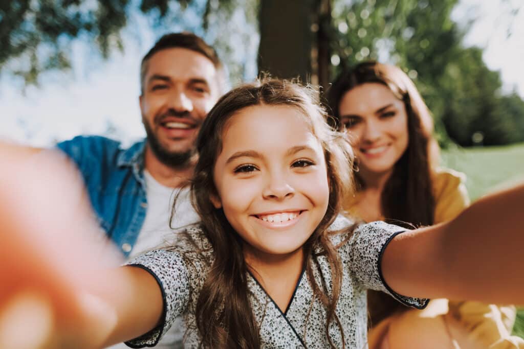 A smiling girl takes a selfie with two adults seated behind her outdoors. The girl wears a patterned shirt and has long hair, while the adults are dressed casually. Green trees and grass are visible in the background.