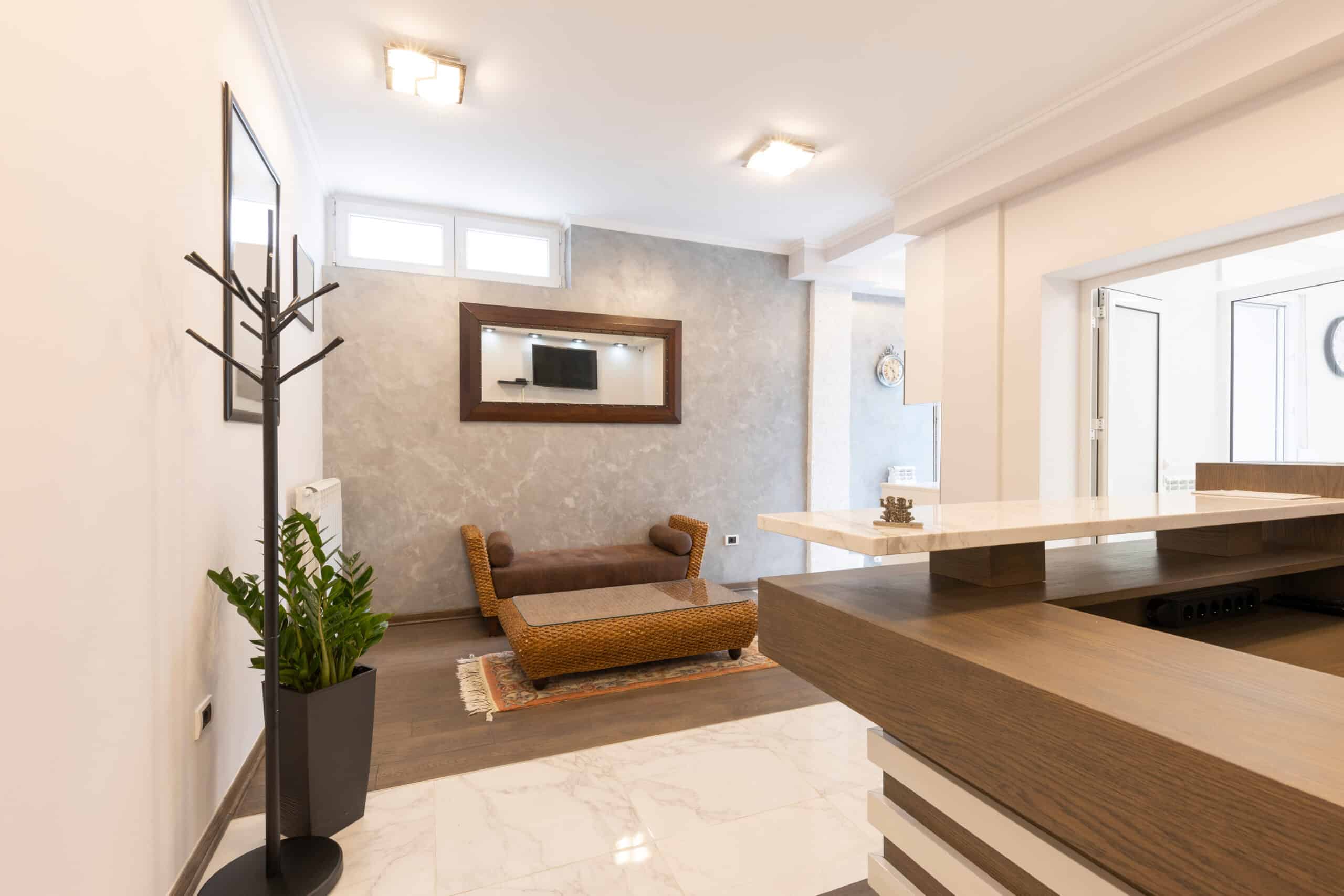 An interior view of a modern living space featuring a brown woven sofa placed on a rug. A sleek wooden counter is in the foreground, with a green plant in a black pot beside it. The walls are light-colored, and there is a wall-mounted television in a wooden frame.