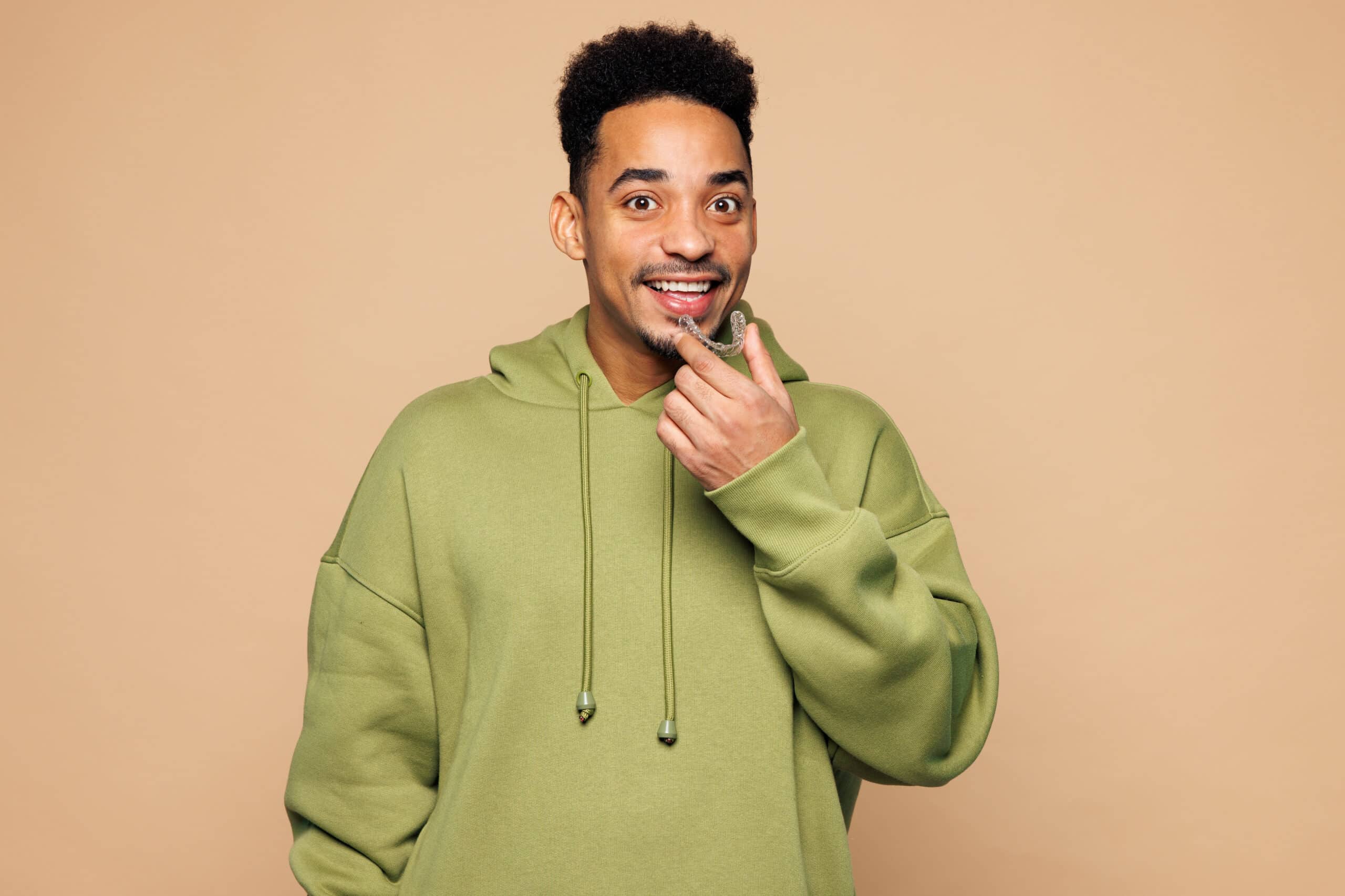 A man in a light green hoodie smiles and points to his chin with his right hand. He has short, curly hair and is set against a neutral beige background.