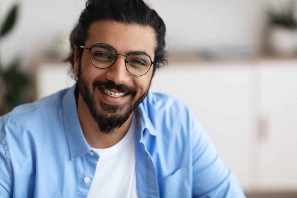 A smiling man with glasses and a beard is sitting indoors, wearing a light blue button-up shirt over a white t-shirt. He has dark, wavy hair tied back and is positioned against a blurred background of a well-lit room with plants and furniture.