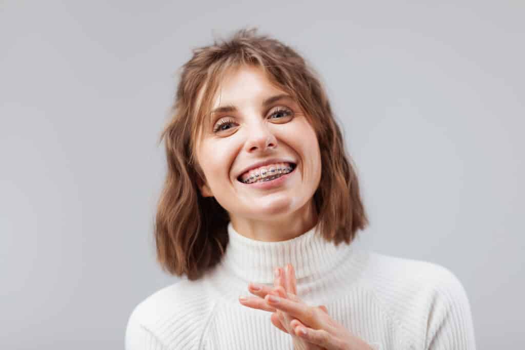 A young woman with shoulder-length hair smiles while wearing braces. She is dressed in a white turtleneck sweater and has her hands in a relaxed position, suggesting a casual and confident demeanor. The background is a soft gray.