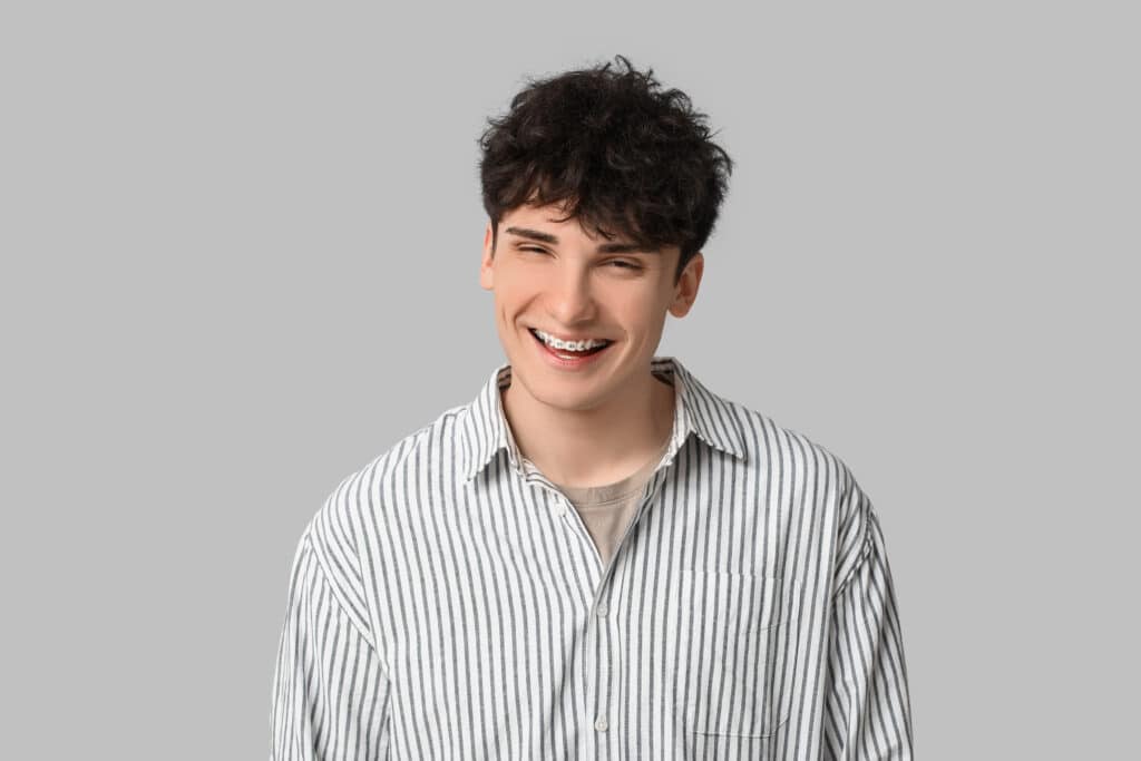 A young man with dark, curly hair and braces is smiling at the camera. He is wearing a light gray and white striped shirt over a beige top. The background is a plain light gray, emphasizing his relaxed expression.
