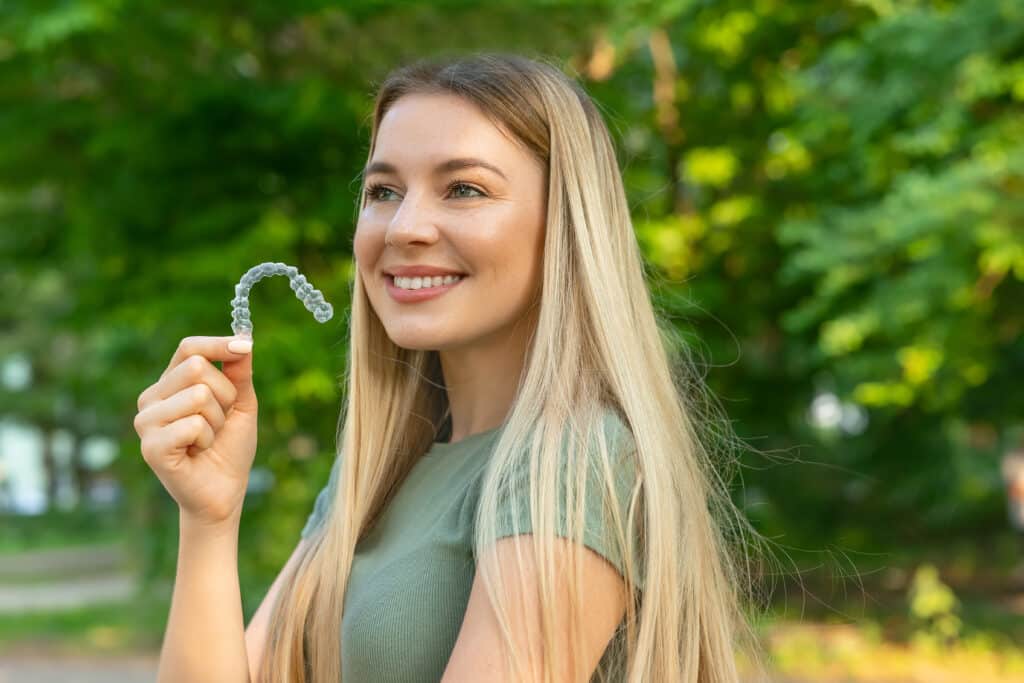 A young woman with long blonde hair holds a clear orthodontic aligner in her right hand, smiling while standing outdoors in a green park. The background features blurred greenery and trees.