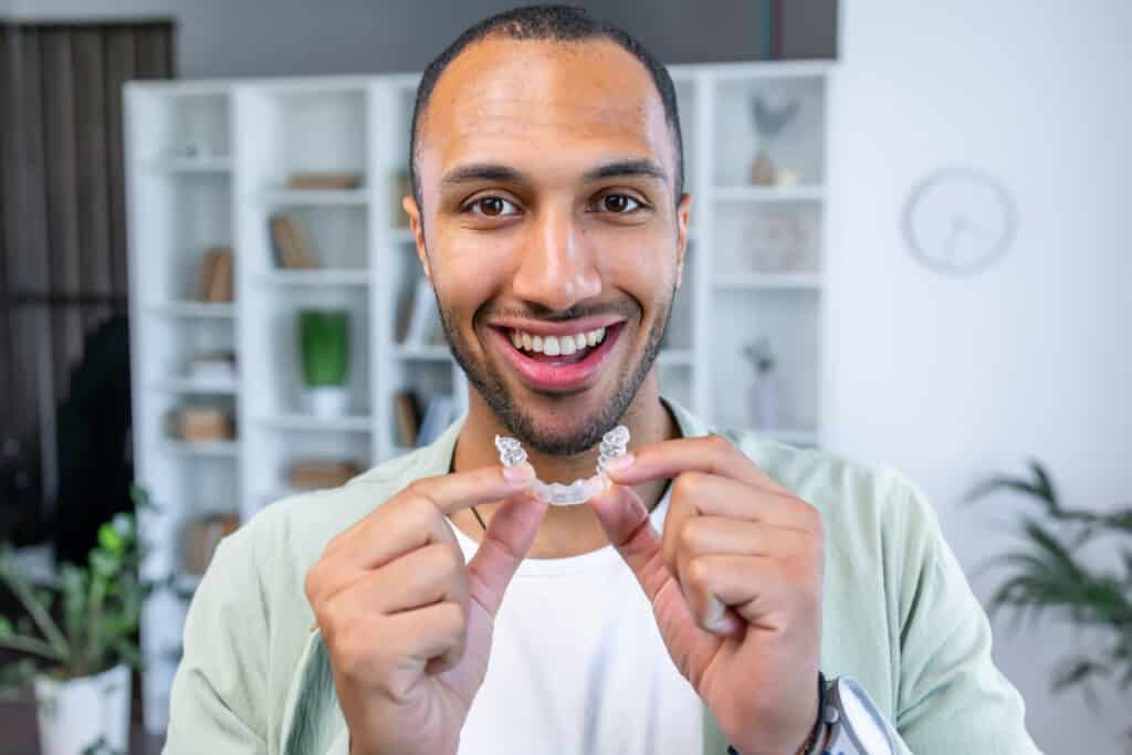 A smiling man holding clear dental aligners, showcasing them between his fingers. He is dressed in a casual outfit and stands in a modern workspace with shelves in the background, featuring plants and decorative items.