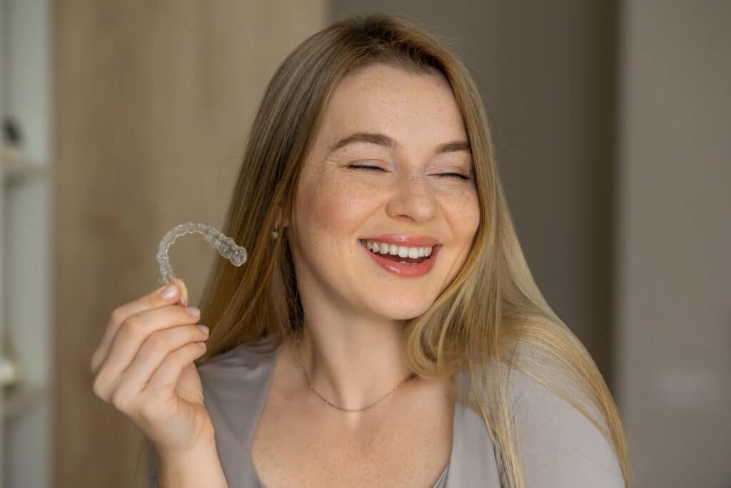 A young woman smiles while holding a clear dental aligner in her right hand. She has long, straight hair and freckles, wearing a light gray top. The background is softly blurred, focusing on her expression and the aligner.