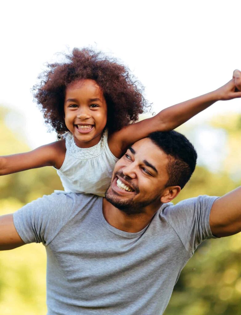 A father holds his daughter, who is smiling widely with her arms outstretched, as she enjoys being lifted in the air. They are outdoors in a green space with blurred trees in the background, indicating a sunny day.