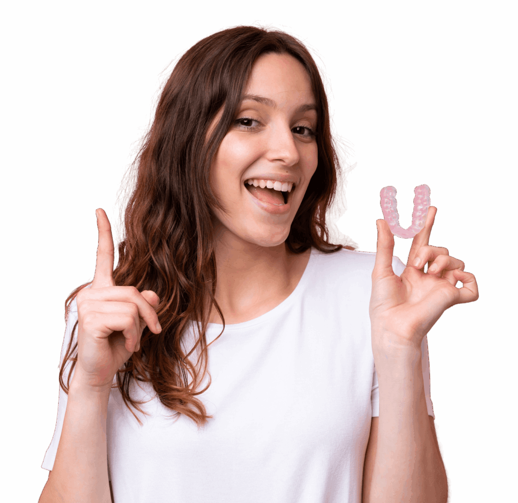 A woman with long brown hair smiles and points up with her left hand. In her right hand, she holds a dental aligner, which is semi-transparent and shaped like a letter 'U.' She is wearing a plain white t-shirt, standing against a plain background.
