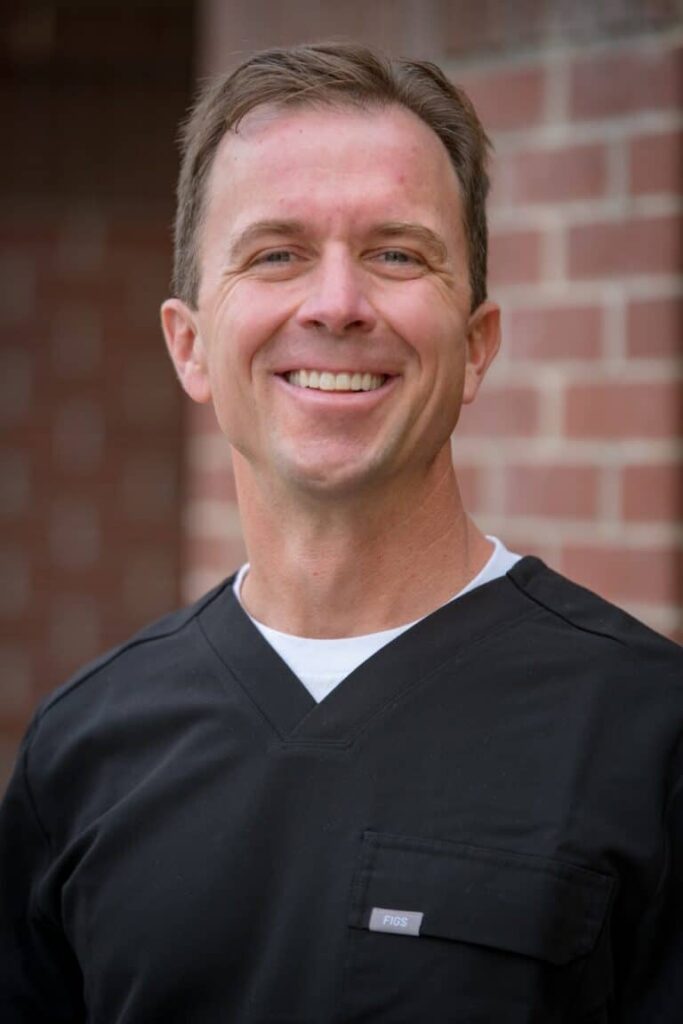 A man in a black scrub top smiles while standing outdoors against a backdrop of a brick wall. He has short brown hair and is wearing a white undershirt. The image conveys a professional appearance.