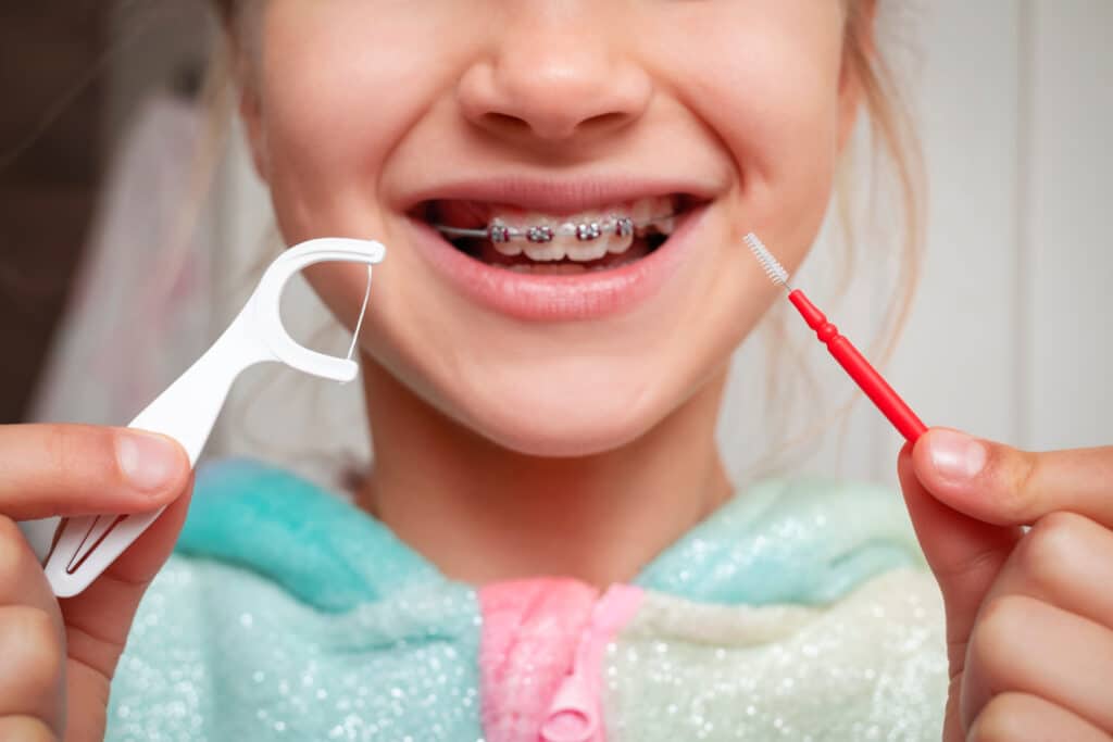 A girl with braces on her teeth is smiling and holding dental care tools: a white floss pick in her left hand and a red interproximal brush in her right hand. She is wearing a colorful, patterned hoodie.