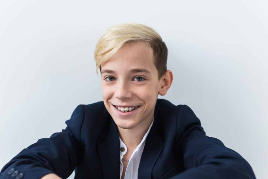 A young boy wearing a dark suit jacket and a white shirt smiles at the camera. He has light blonde hair and is seated against a plain white background, with his arms resting on his knees. The image conveys a friendly and approachable demeanor.