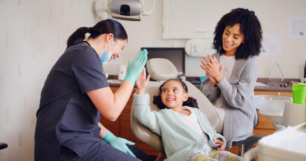 A child sitting in a dental chair smiles and high-fives a dental hygienist wearing gloves and a mask. A parent sits nearby, clapping and smiling. The dental office has a warm, welcoming atmosphere with medical equipment visible in the background.
