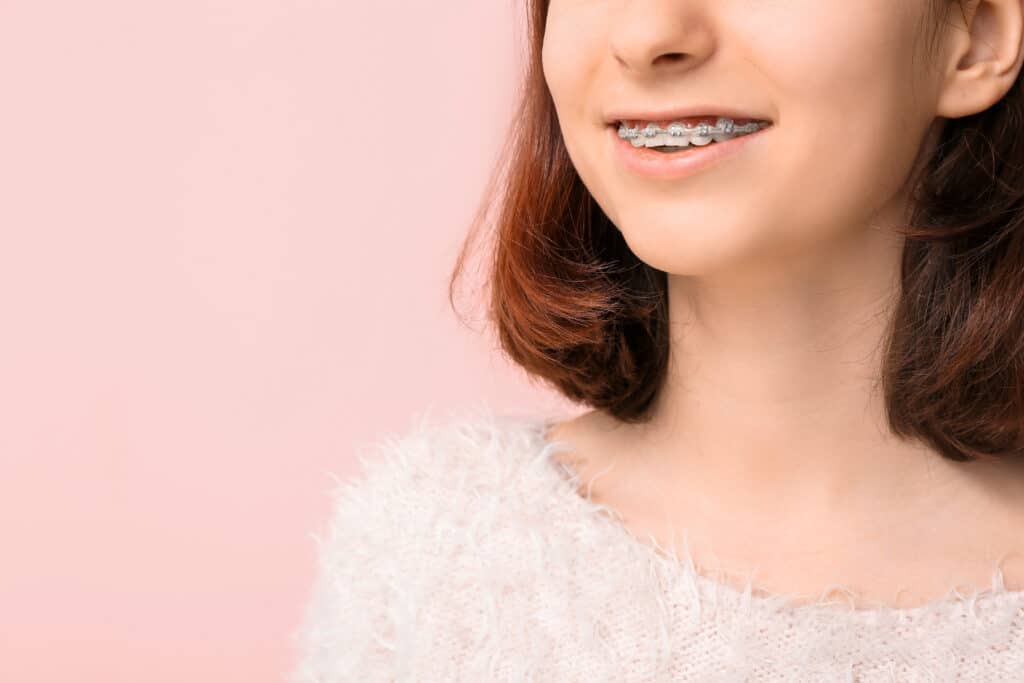 Close-up of a young woman smiling, showing braces on her teeth. She has shoulder-length, brown hair, and is wearing a light-colored, fluffy sweater. The background is a soft pink color.