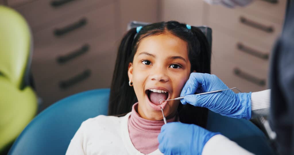 A young girl in a dental chair smiling while a dentist, wearing blue gloves, examines her teeth with a dental tool. The background shows dental equipment and furniture, creating a clinical environment.