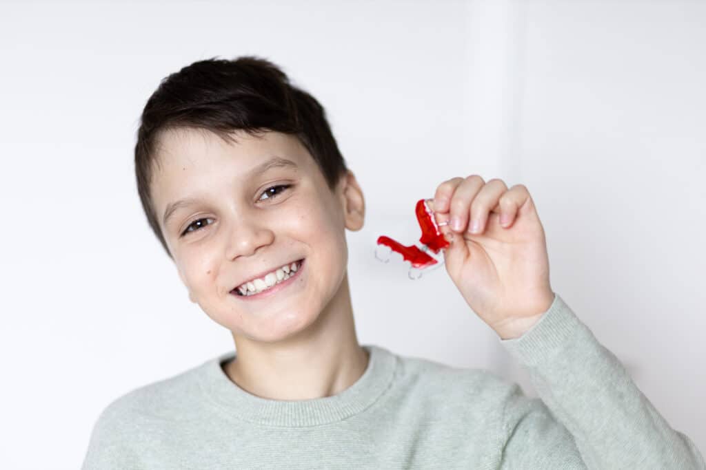 A smiling boy holding a piece of red candy in his hand, wearing a light gray sweater. He has short brown hair and is standing against a light background.