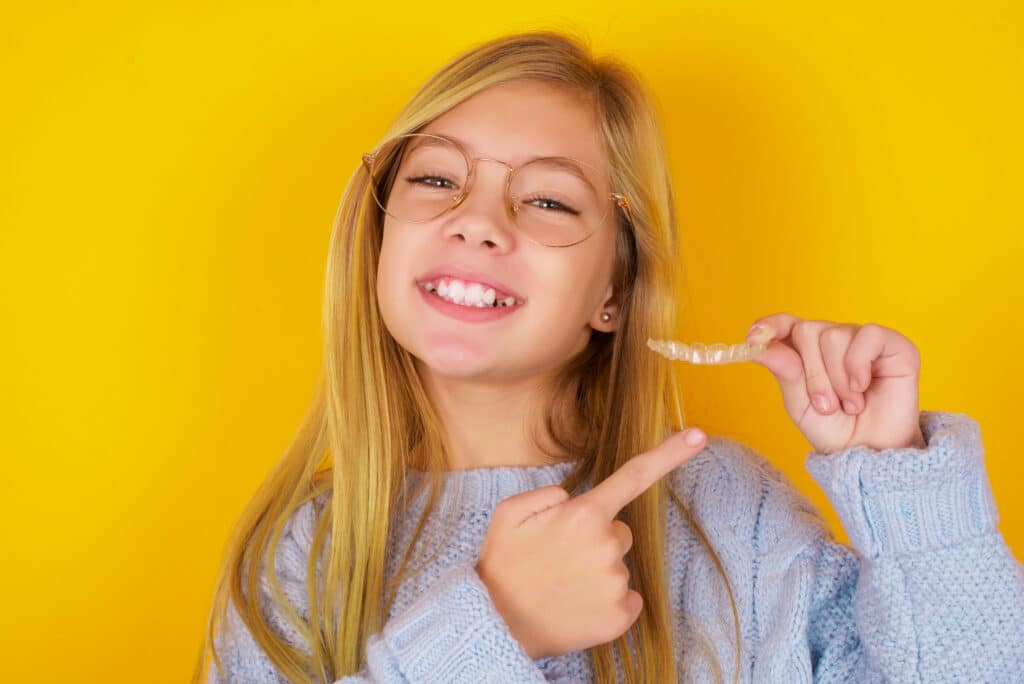 A smiling girl with long blonde hair and glasses points to a dental retainer in her hand. She is wearing a light blue sweater against a bright yellow background.