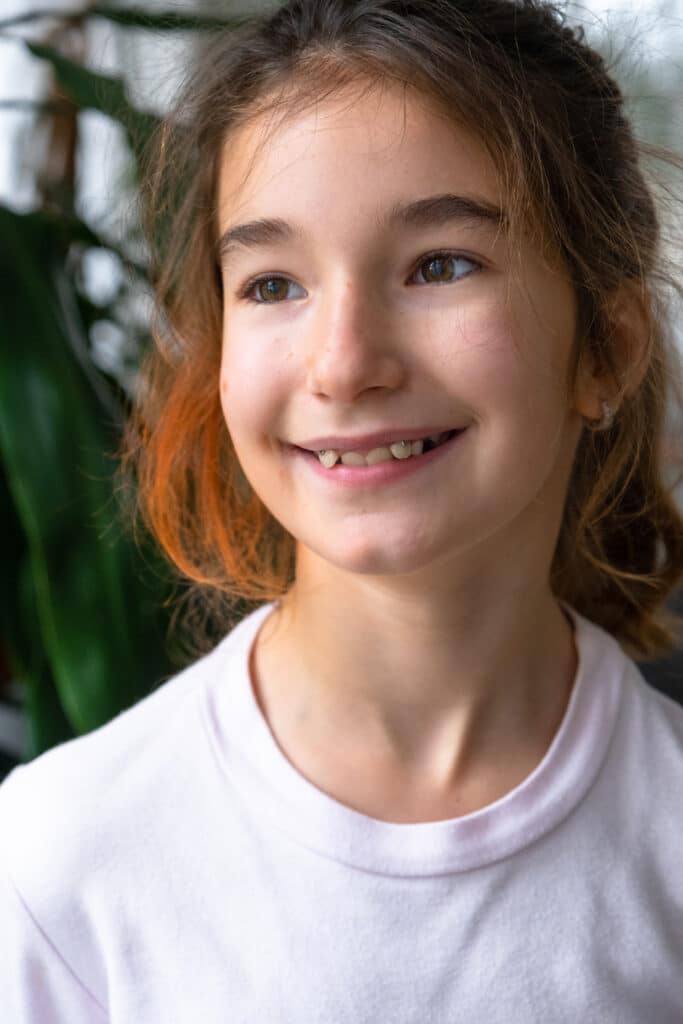 A young girl with brown hair and a slight smile is shown in a close-up portrait. She is wearing a light pink shirt and is against a backdrop of green plants, creating a natural and relaxed atmosphere.