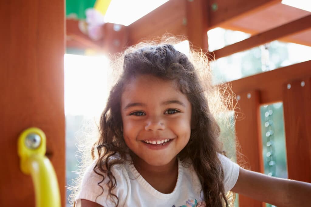 A smiling girl with curly hair is sitting on a wooden playground structure. Sunlight filters through, illuminating her face. She wears a white t-shirt and is positioned near a yellow handle, surrounded by wooden elements of the playground.