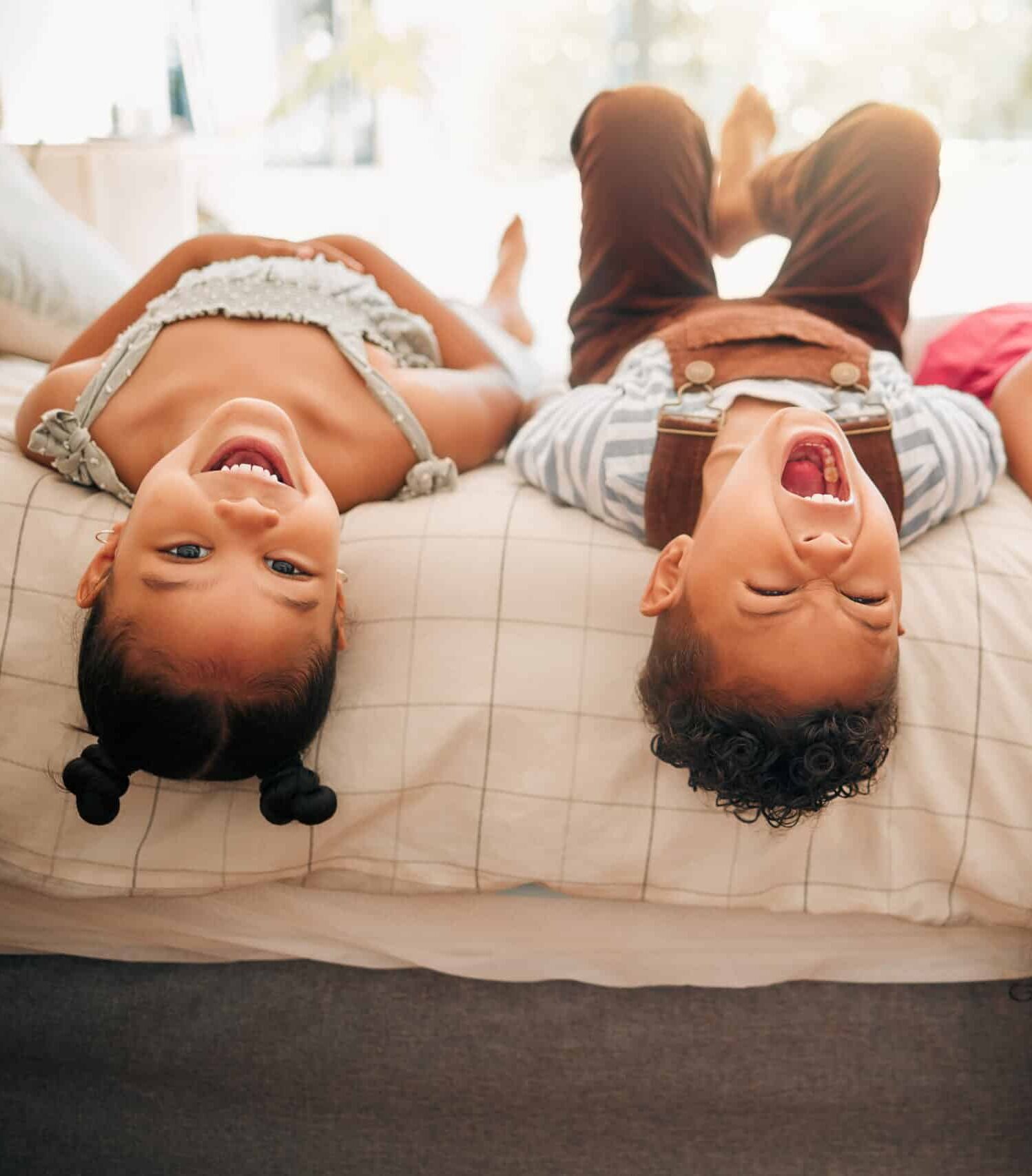 Three children laughing while laying on a bed, with their heads near the edge and legs in the air. The girl on the left wears a light gray outfit, the boy in the center has on a striped shirt and brown overalls, and the girl on the right is dressed in a pink top.
