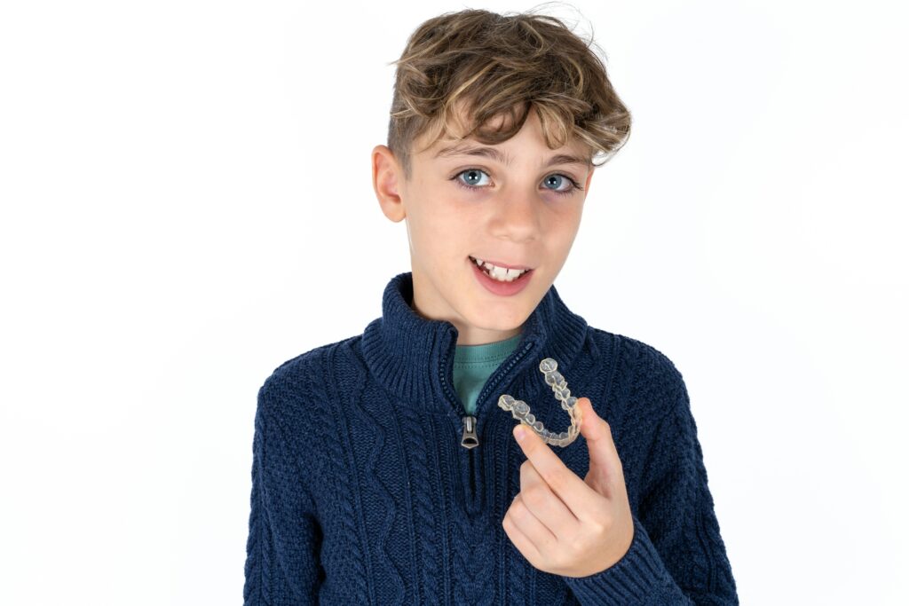 A young boy with light brown hair smiles while holding a clear dental aligner in his right hand. He is wearing a navy blue sweater and has a green shirt underneath. The background is plain white.