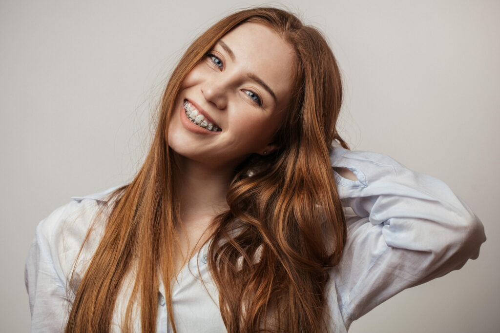 A young woman with long, reddish-brown hair smiles while posing against a plain background. She is wearing a light blue button-up shirt and has braces on her teeth, her right hand resting behind her head in a relaxed manner.