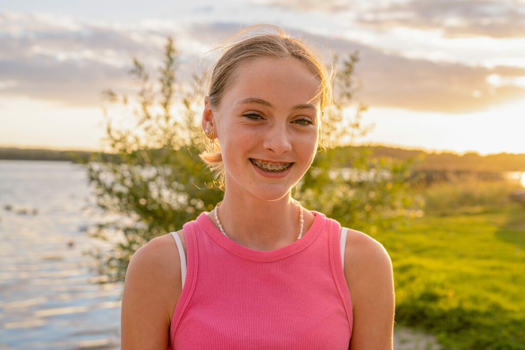 A young girl with braces smiling in front of a lake during sunset. She is wearing a pink sleeveless shirt, and her hair is styled casually. The background features a body of water and greenery, with soft sunlight illuminating the scene.