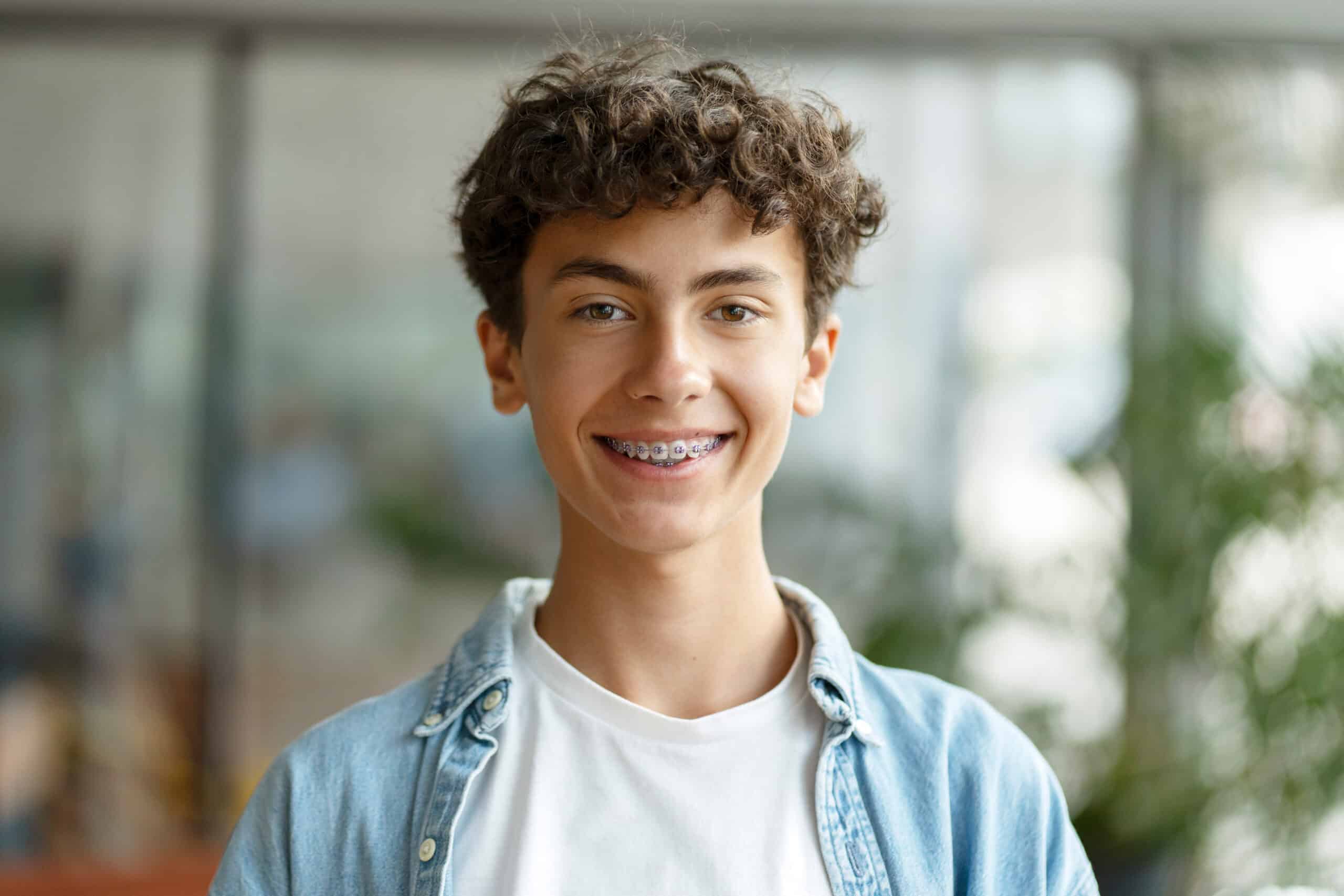 A teenager with curly hair and braces stands smiling in a bright indoor space. He wears a white t-shirt under a denim jacket. Blurred background features greenery and soft light, suggesting a casual environment.
