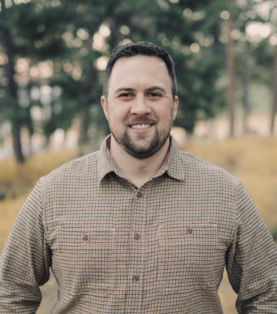 A man with short dark hair and a trimmed beard stands outdoors, wearing a brown checkered shirt. Behind him, there are trees and dry grass, suggesting a natural setting. The lighting indicates either morning or evening.